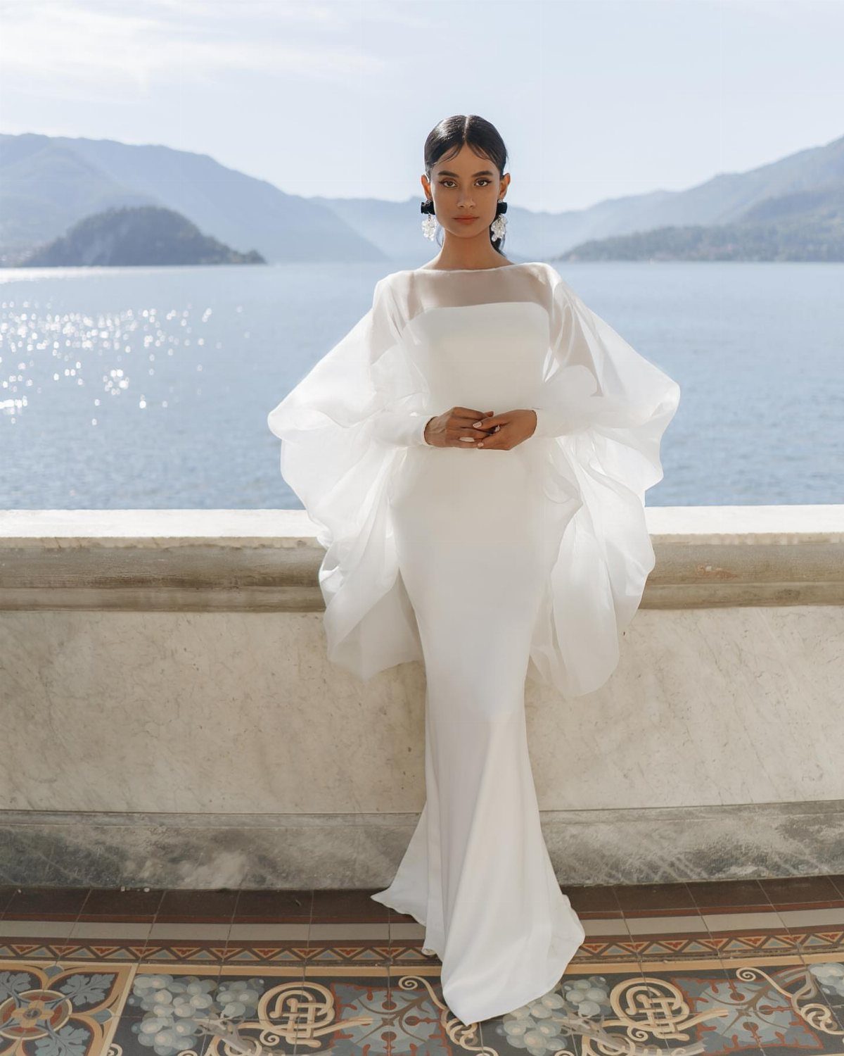 Woman in a white wedding gown with a sheer, voluminous white cape by a lake