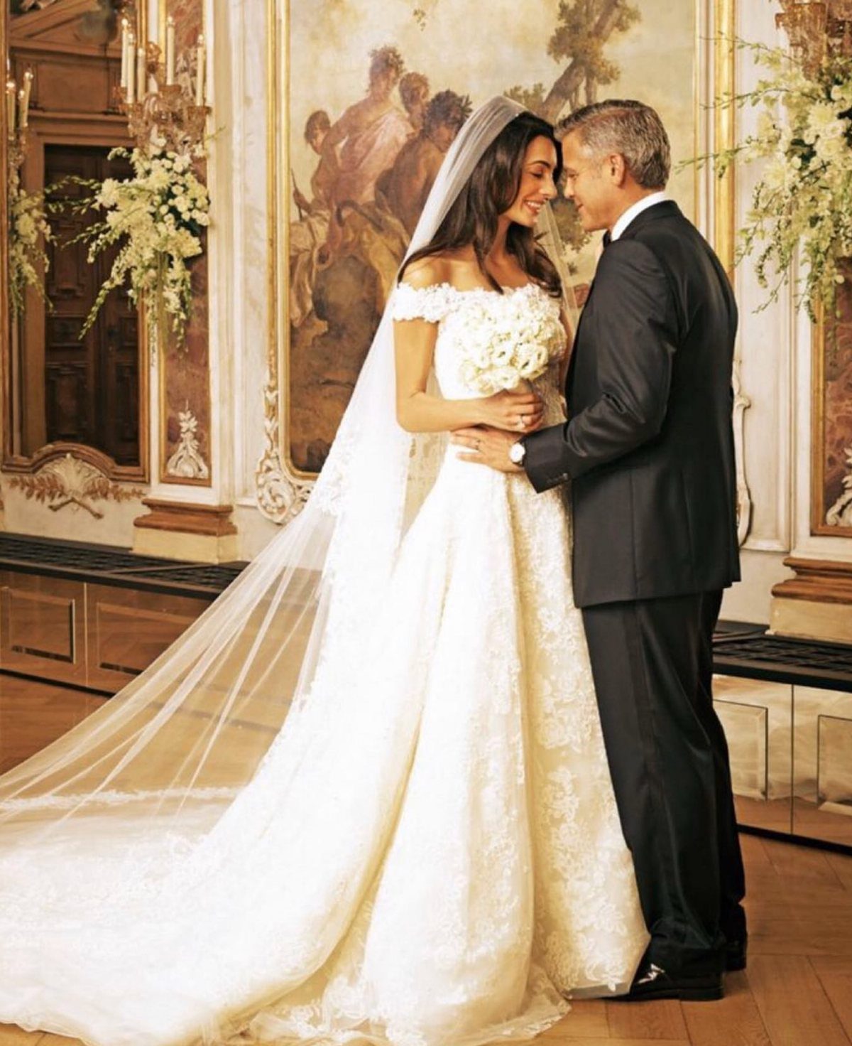 Amal Clooney in an off-the-shoulder lace wedding gown standing with George Clooney.