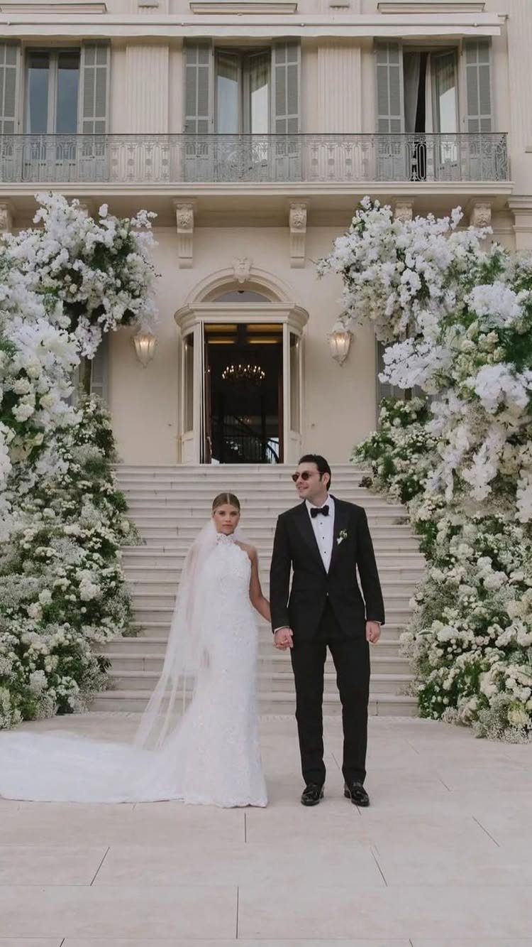 Bride in white lace gown and groom in tuxedo before floral estate entrance