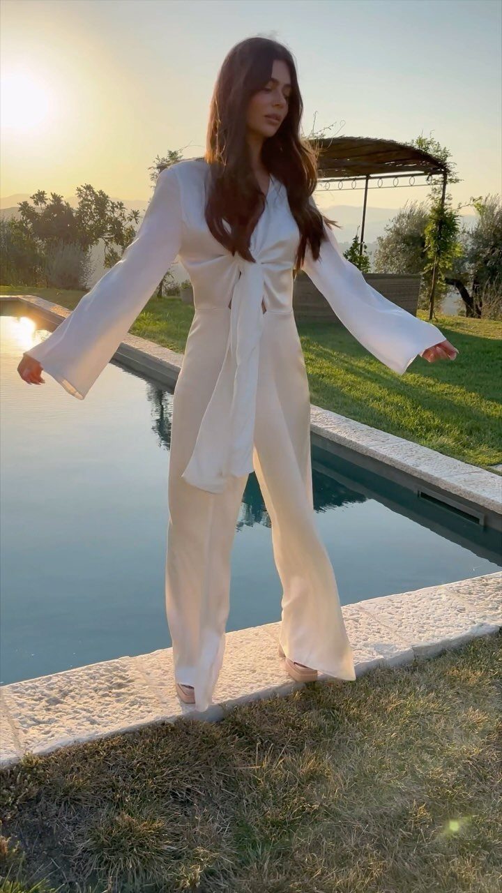 Woman in a white wide-leg bridal jumpsuit standing by a pool at sunset