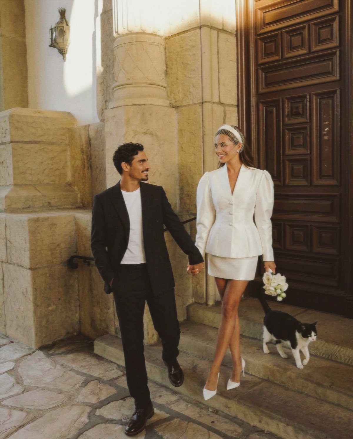 Woman in white cropped jacket and matching skirt holding hands with man in suit.