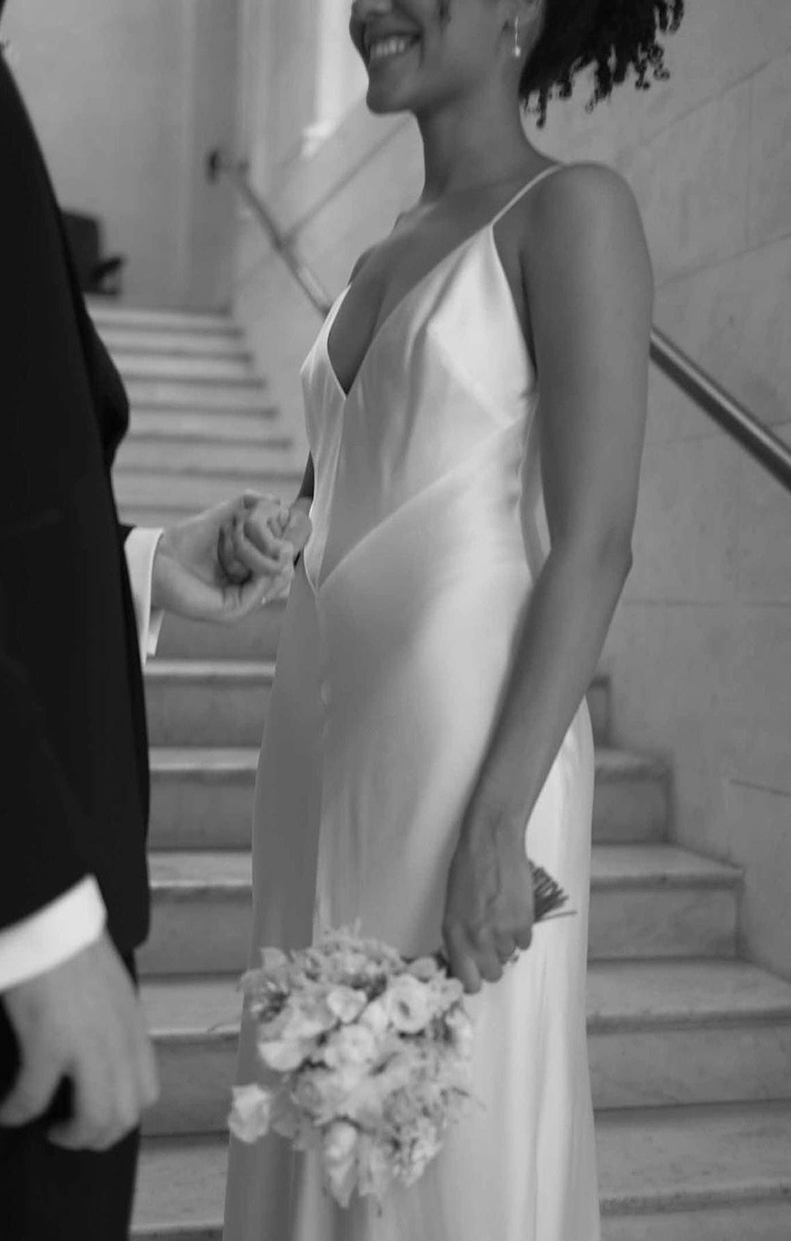 Woman in a white silk slip dress holding a bouquet on stone stairs.
