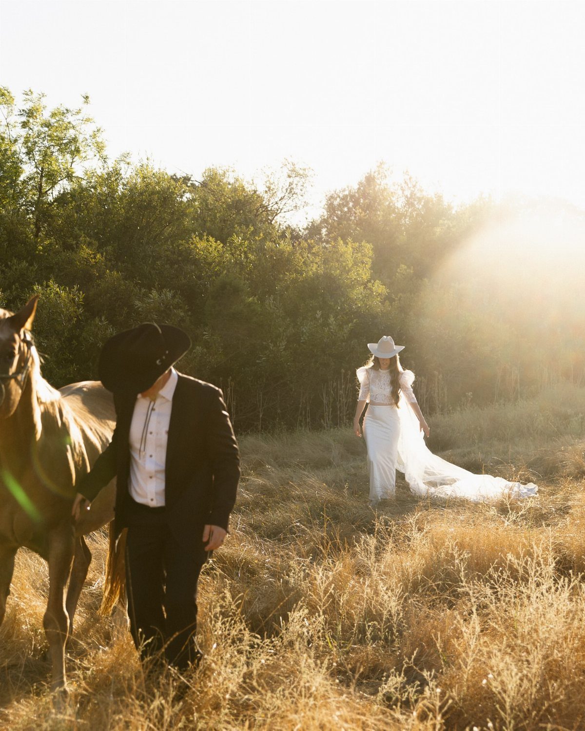 Bride in white dress and cowboy hat standing in a golden field with horse