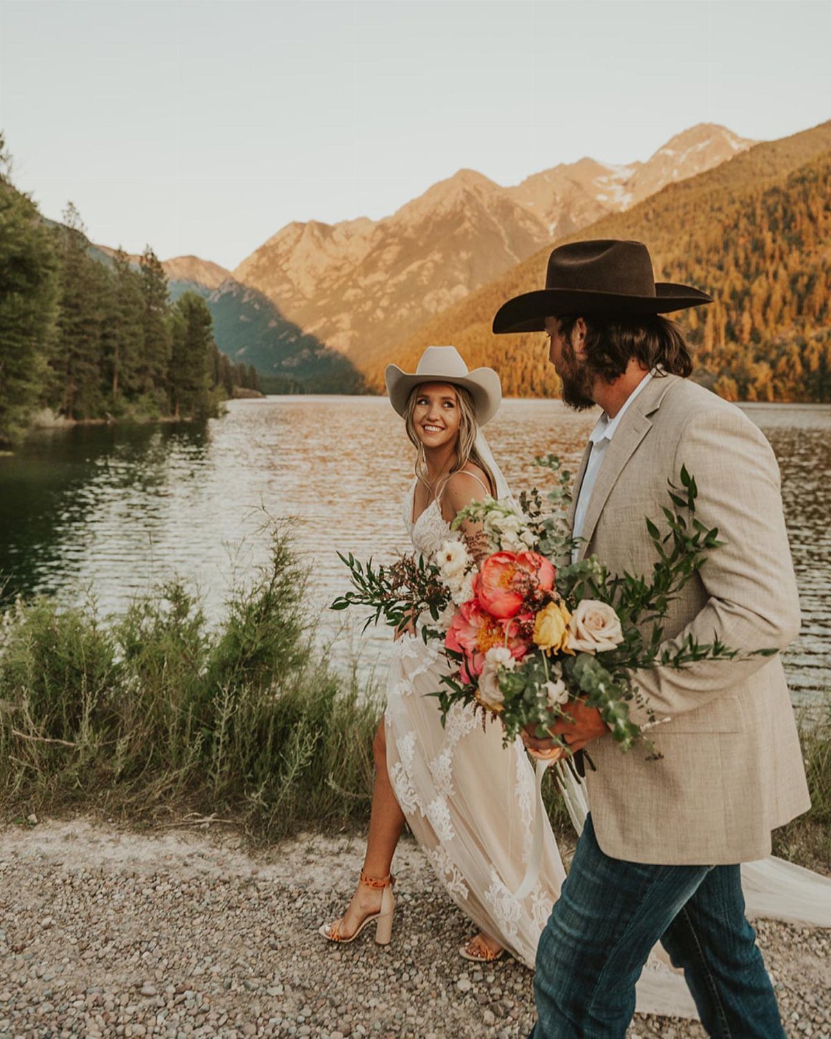Bride in white lace dress and cowboy hat walking by a mountain lake