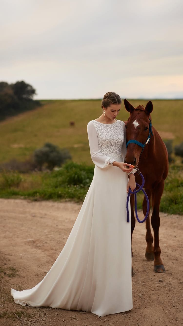 Woman in a white lace wedding dress standing with a brown horse outdoors