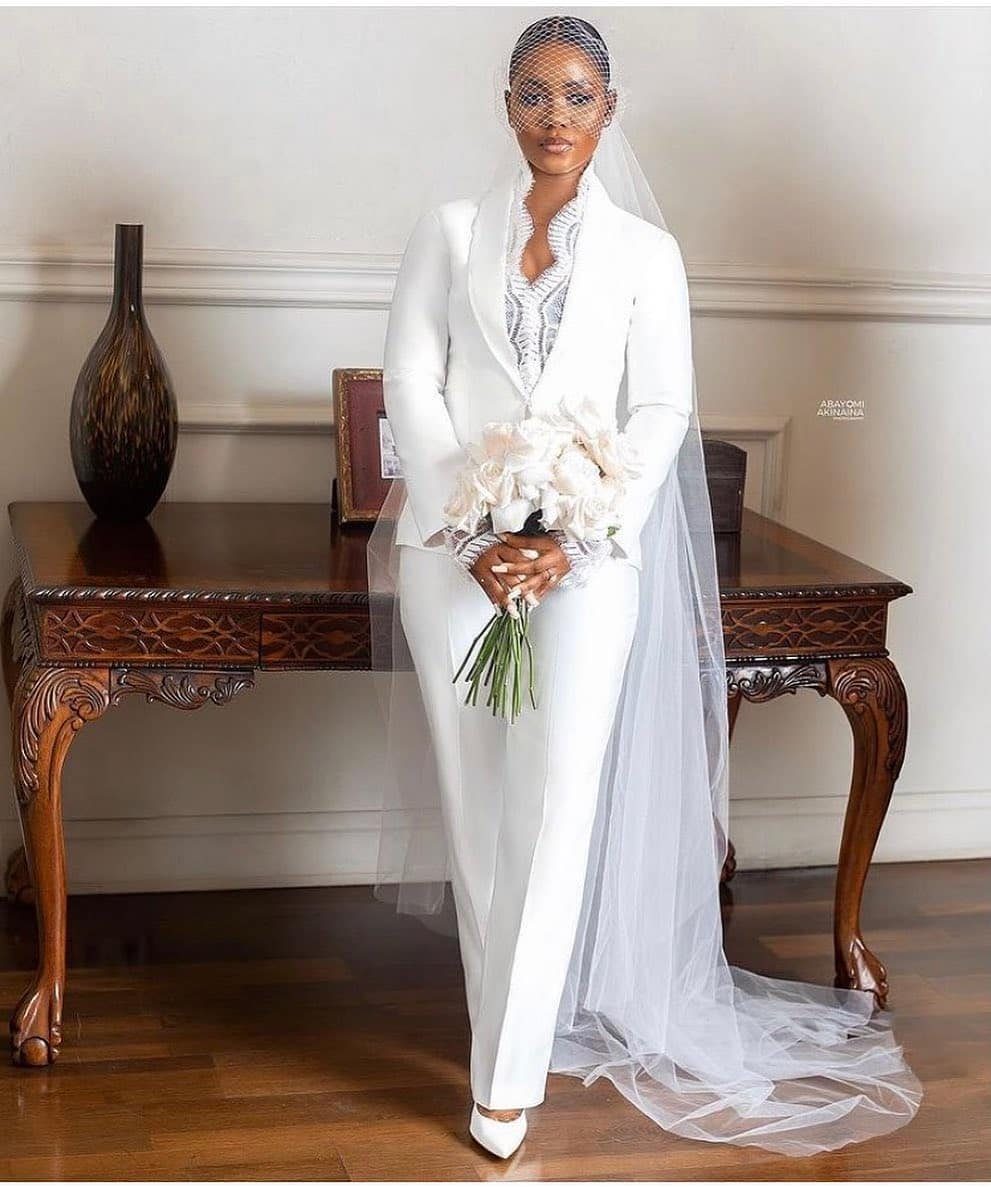 Woman in a white bridal pantsuit with a long veil holding white flowers.