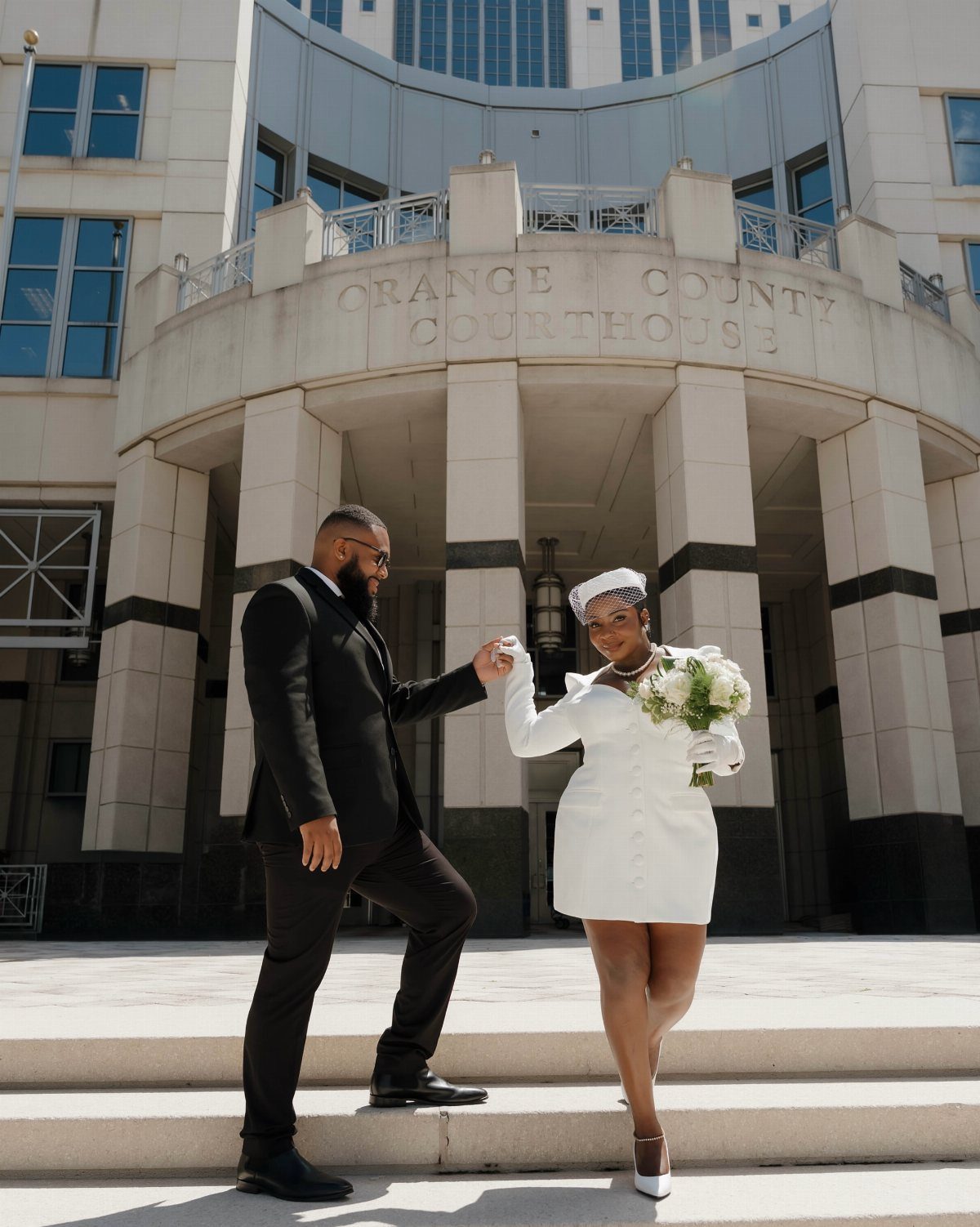 Couple in wedding attire posing on the steps of a stone courthouse building.