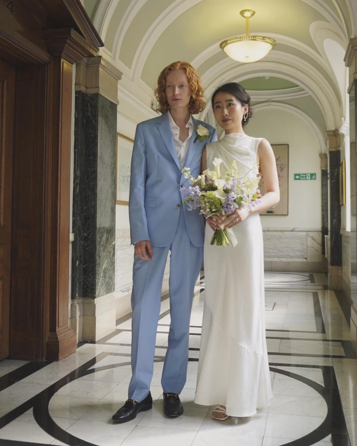 Couple in a marble hallway, man in light blue suit, woman in white gown.