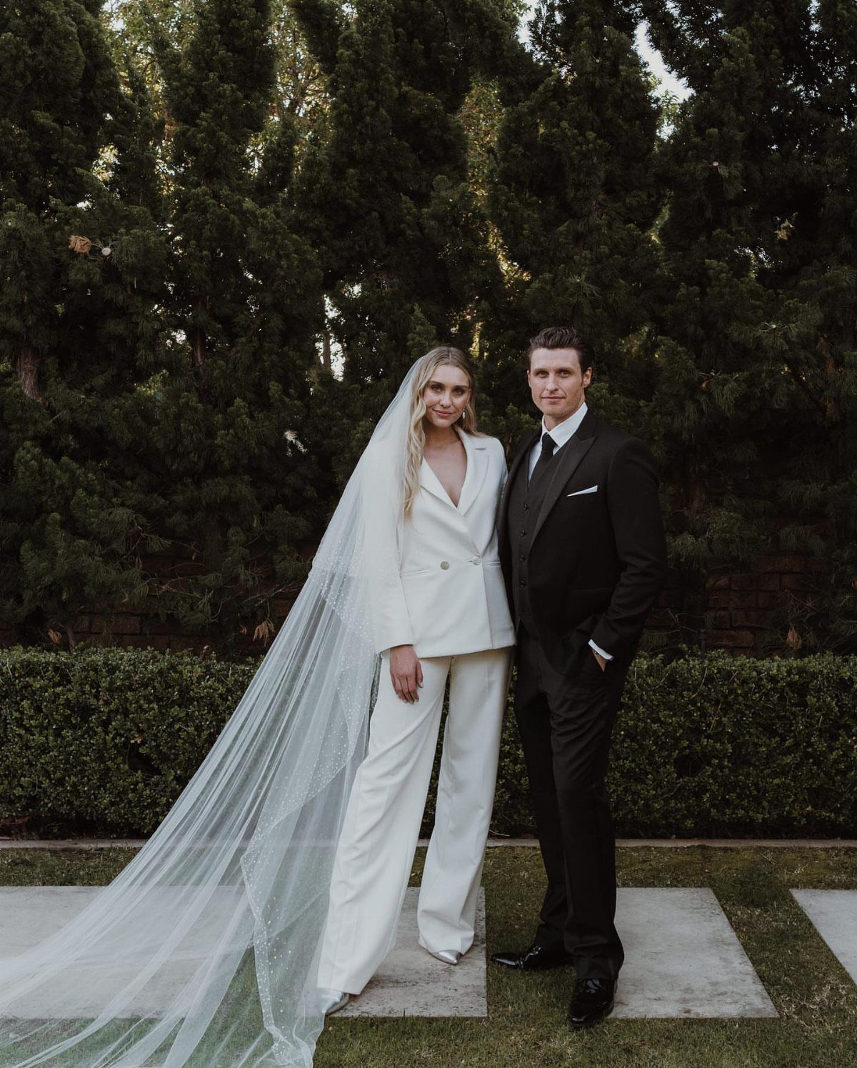 Bride in a white tailored suit and veil standing with groom in black suit.