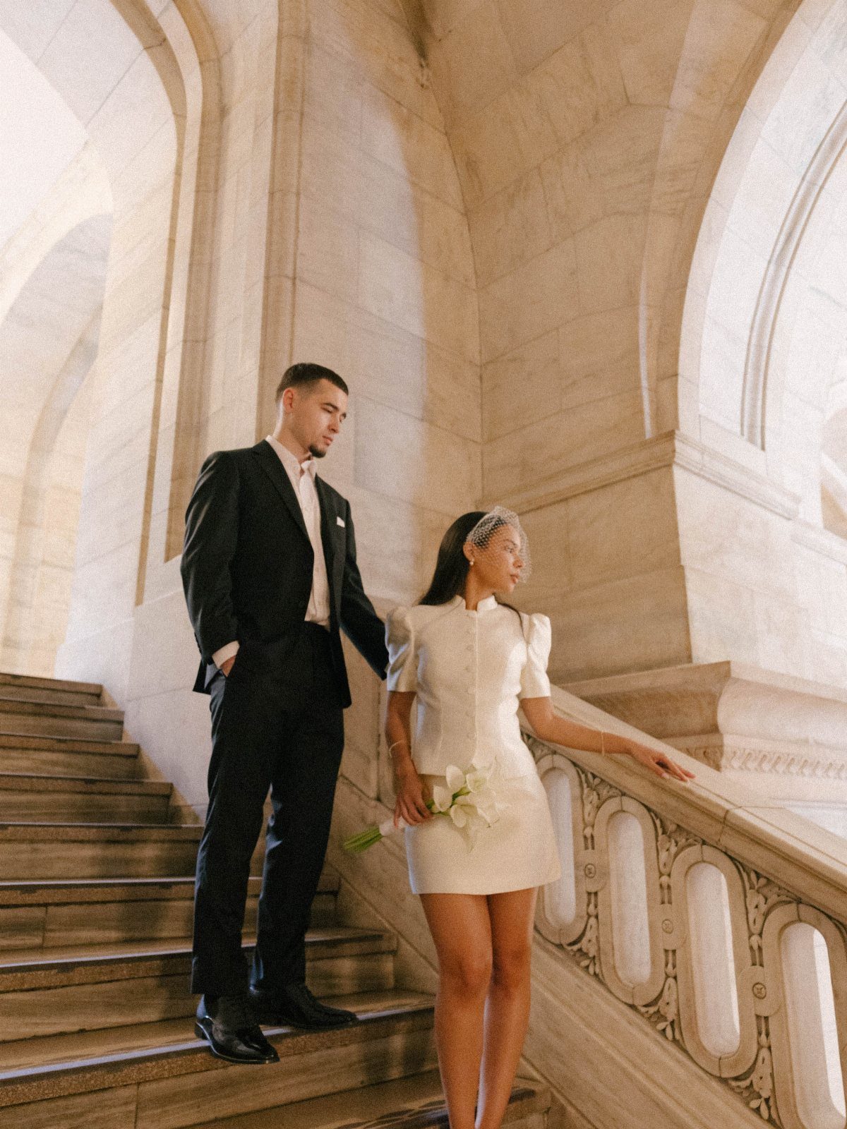 Couple on stone stairs, woman wearing a short structured white wedding dress.