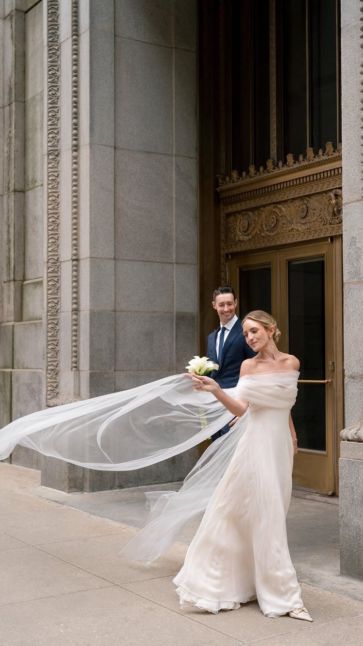Bride in white off-the-shoulder dress and groom in blue suit outside stone building