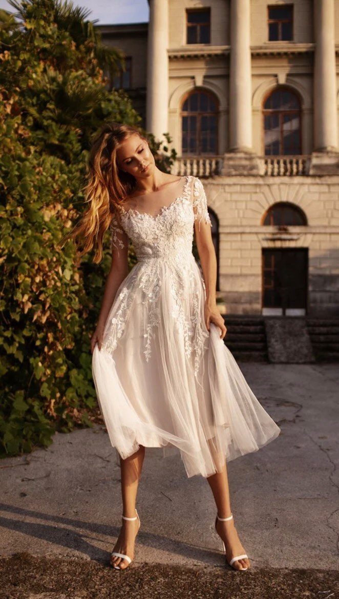 Woman in a white lace tea-length wedding dress standing before a classical building.