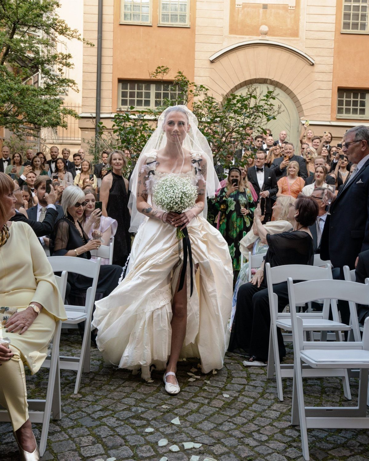 Bride in an avant-garde white wedding gown walking down a cobblestone aisle.