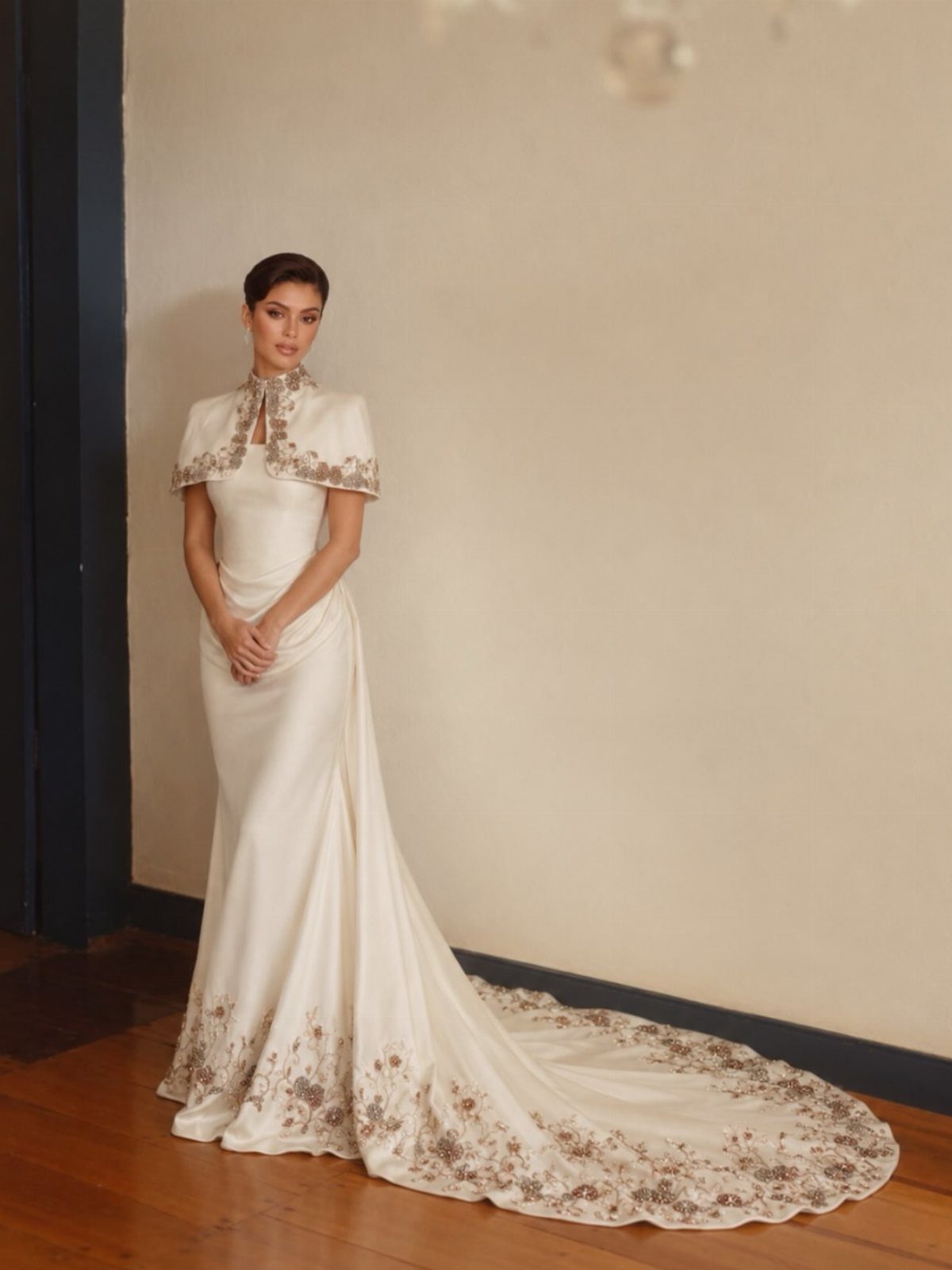 Woman in a sleek white bridal gown with gold floral embroidery and capelet.