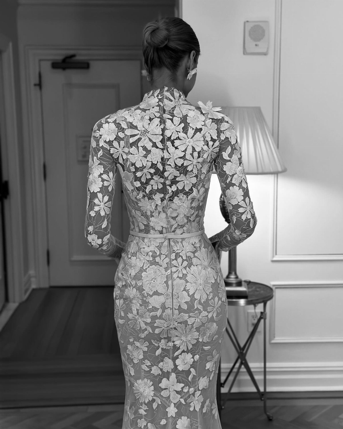 Woman from behind wearing a long-sleeved floral lace bridal gown in a room.