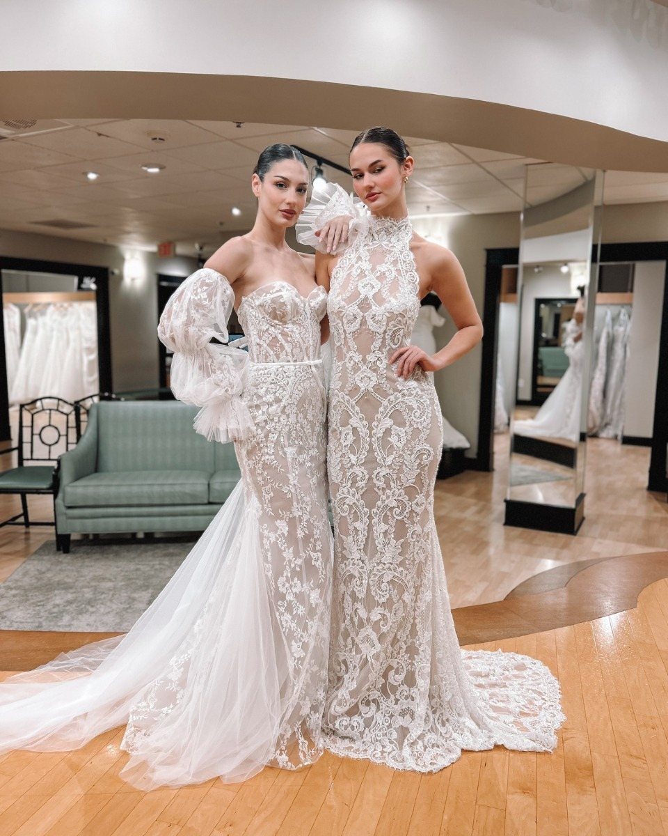 Two women wearing figure-hugging white lace bridal gowns in a wedding boutique.