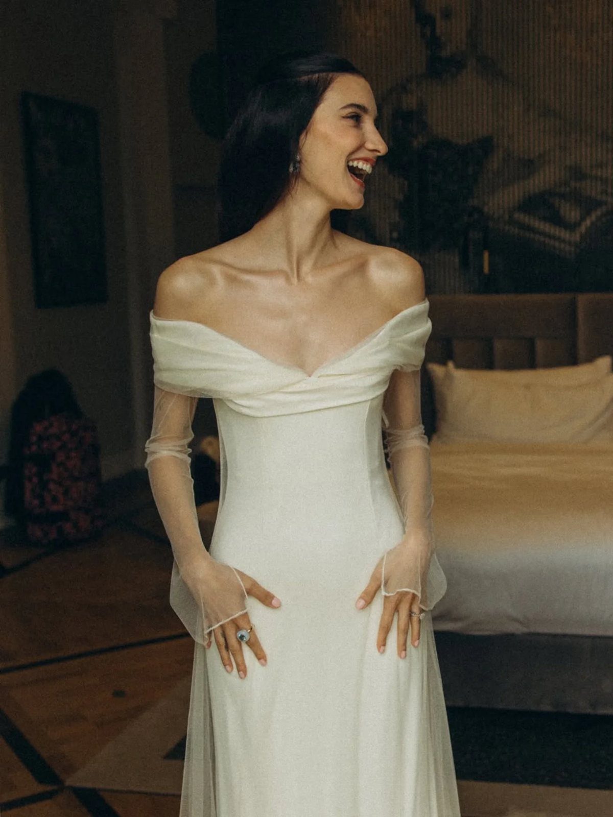 Woman laughing in a modern off-the-shoulder white bridal gown with sheer sleeves.