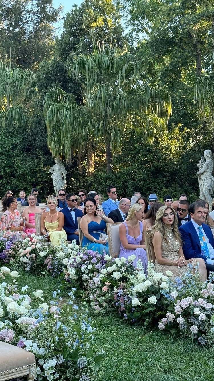 Wedding guests in colorful attire seated in a garden with white floral borders.