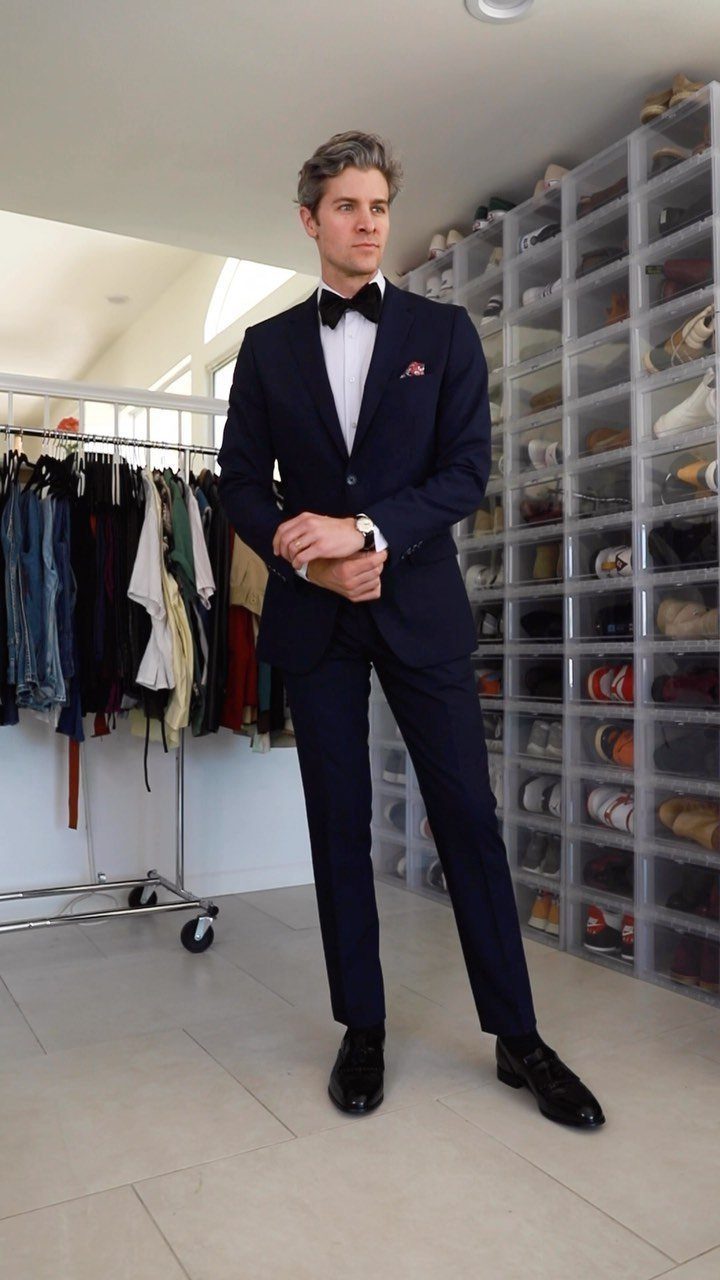 Man in navy blue tuxedo and black bow tie posing in a closet