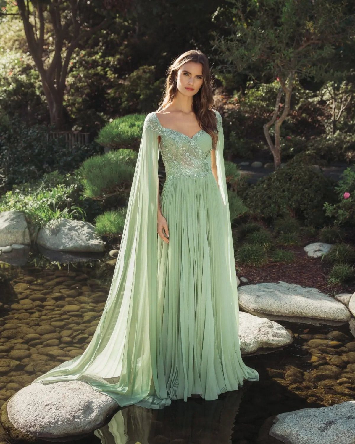 Woman in a sage green flowing dress with cape sleeves in a garden