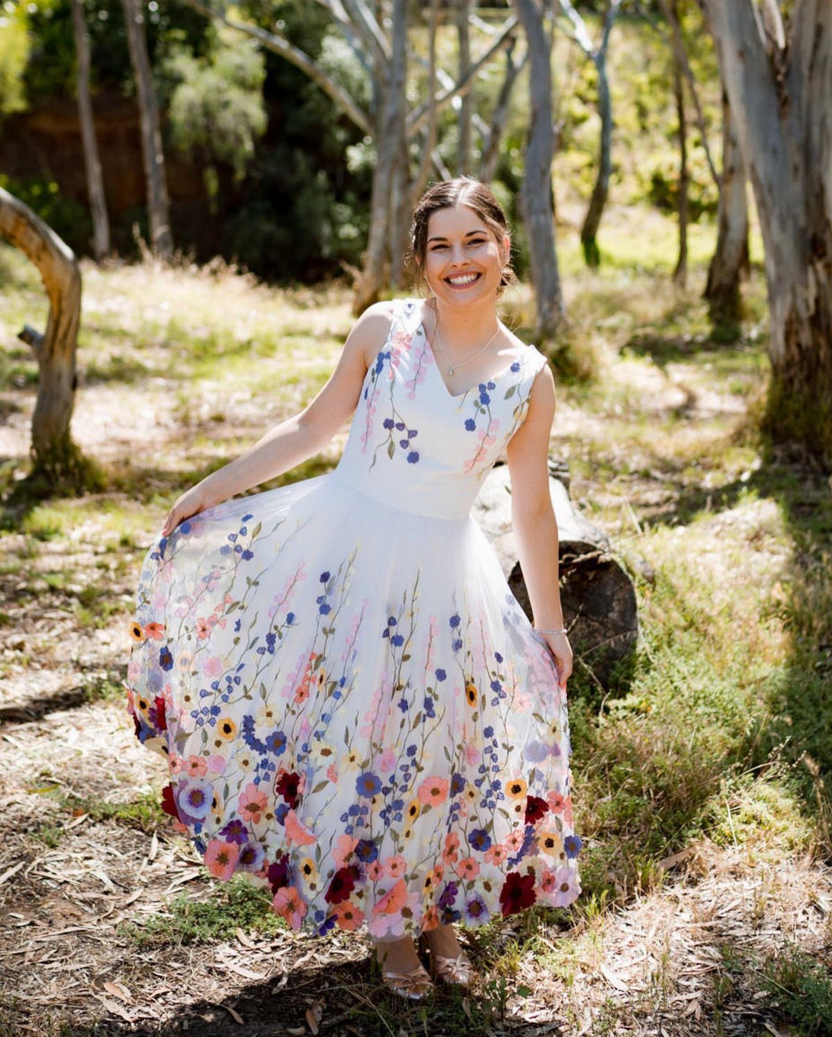 Woman in a white floral wedding dress smiling in a sunny wooded forest