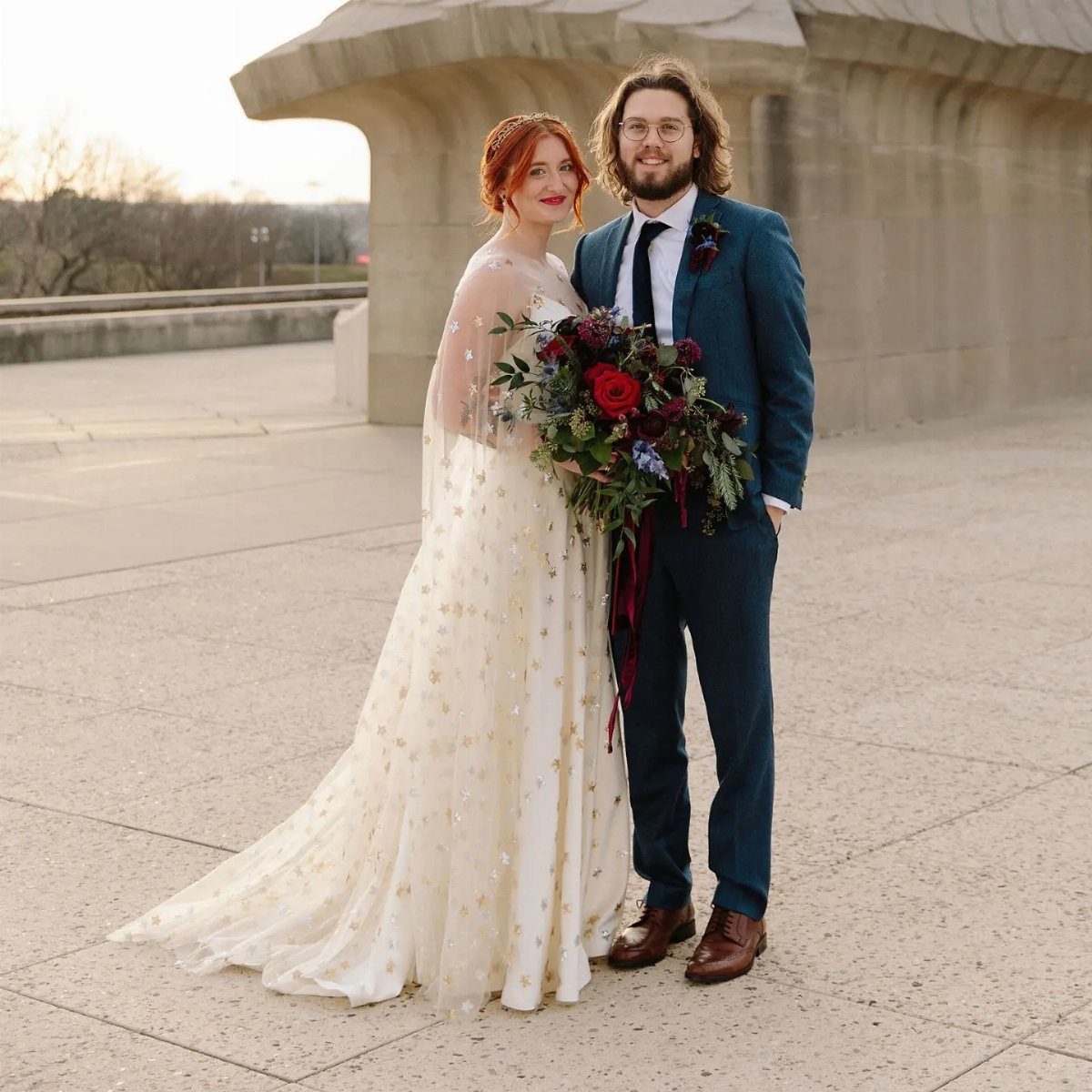 Bride in gold-starred wedding gown and groom in blue suit posing outdoors.