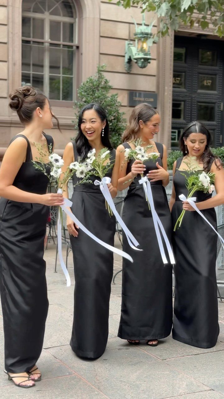 Four bridesmaids in long sleeveless black satin dresses holding white floral bouquets.