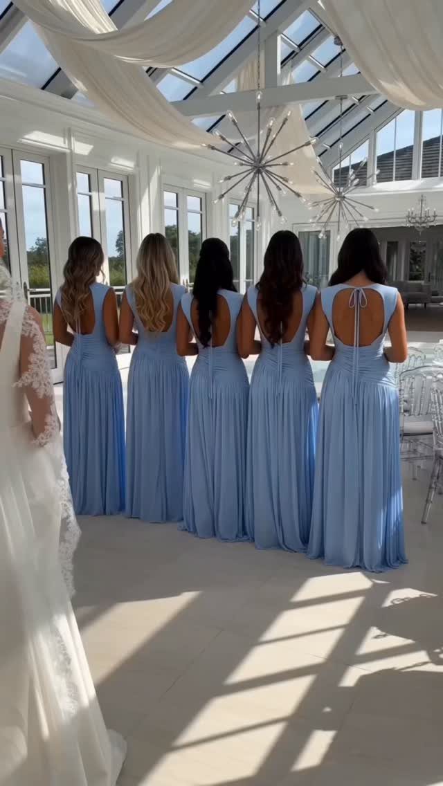 Five bridesmaids in light blue floor-length dresses standing in a bright white sunroom.