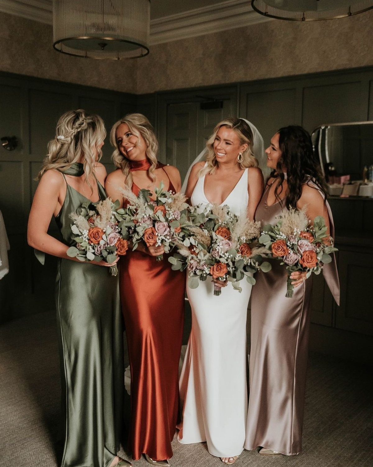 Bride and bridesmaids in autumn-colored satin dresses holding floral bouquets indoors