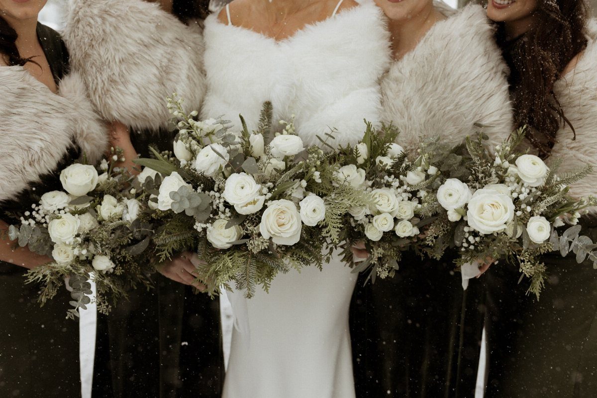 Bride and bridesmaids wearing white faux-fur shawls and holding white floral bouquets.