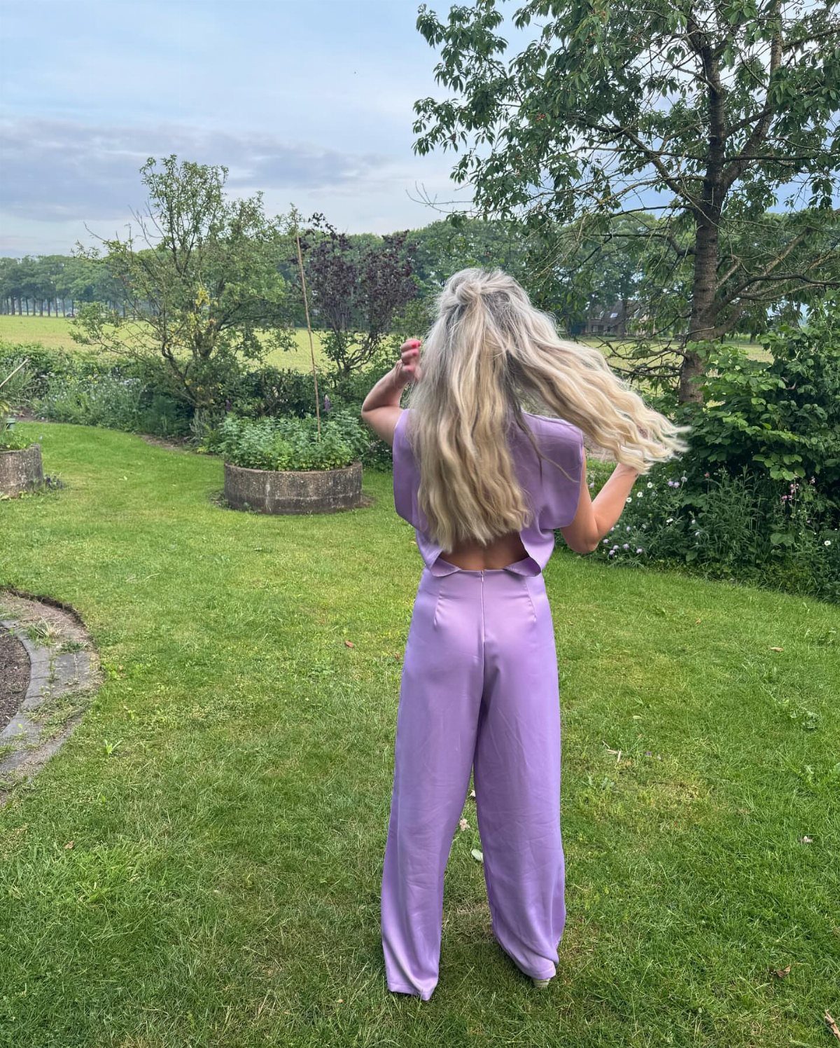 Woman in a purple dressy jumpsuit standing in a green garden setting.