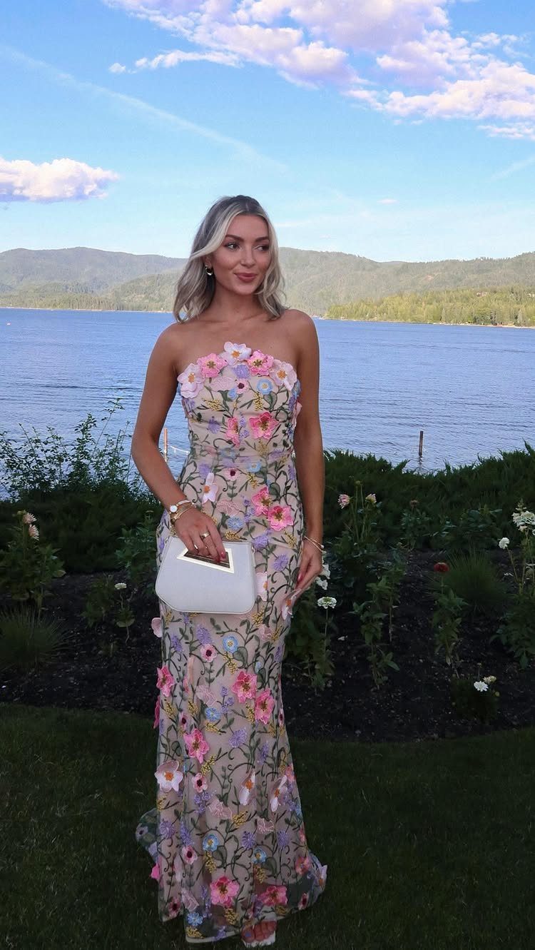 Woman in a strapless floral print gown holding a white handbag by a lake