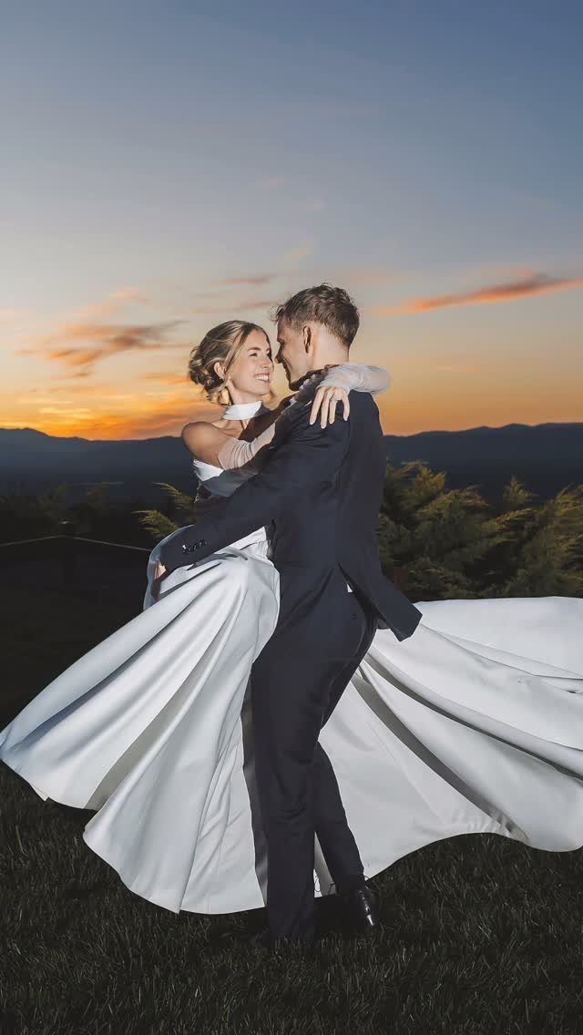 Groom lifting bride in a white gown against a mountain sunset sky.
