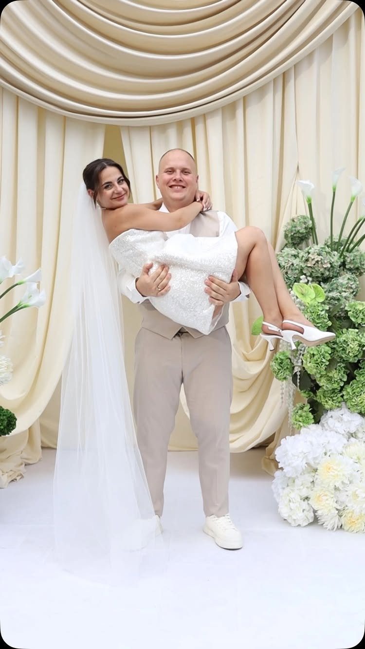 Groom lifting bride in a white wedding dress and veil before cream curtains