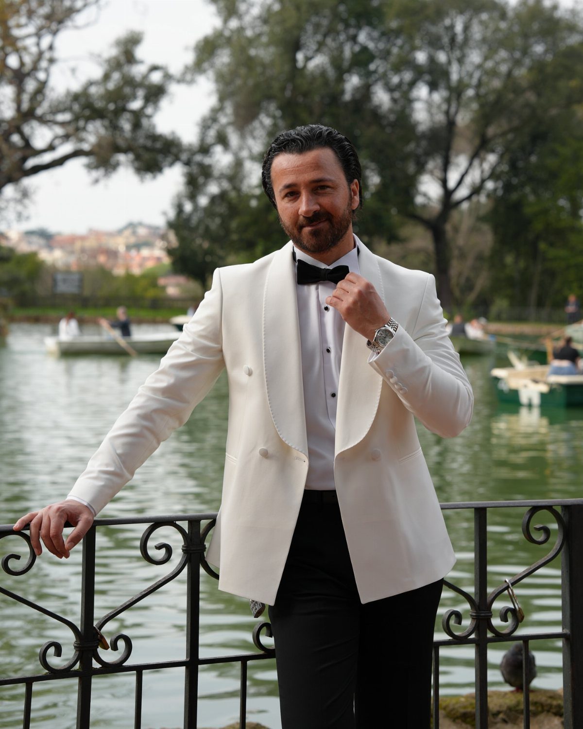 Man in white dinner jacket and black bow tie posing by a lake