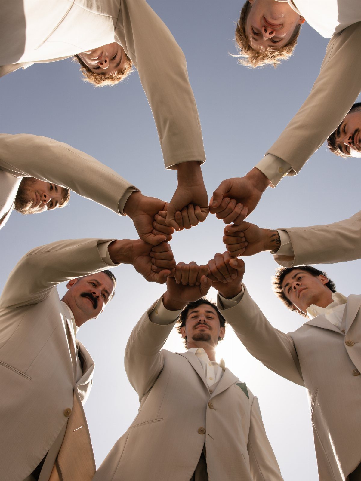 Six men in beige suits joining fists in a huddle against a blue sky.