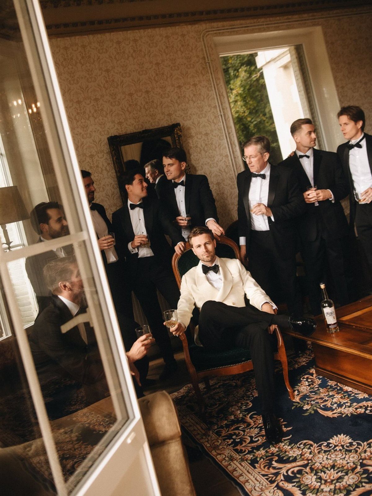 Groom in white tuxedo jacket surrounded by groomsmen in black tuxedos holding champagne.
