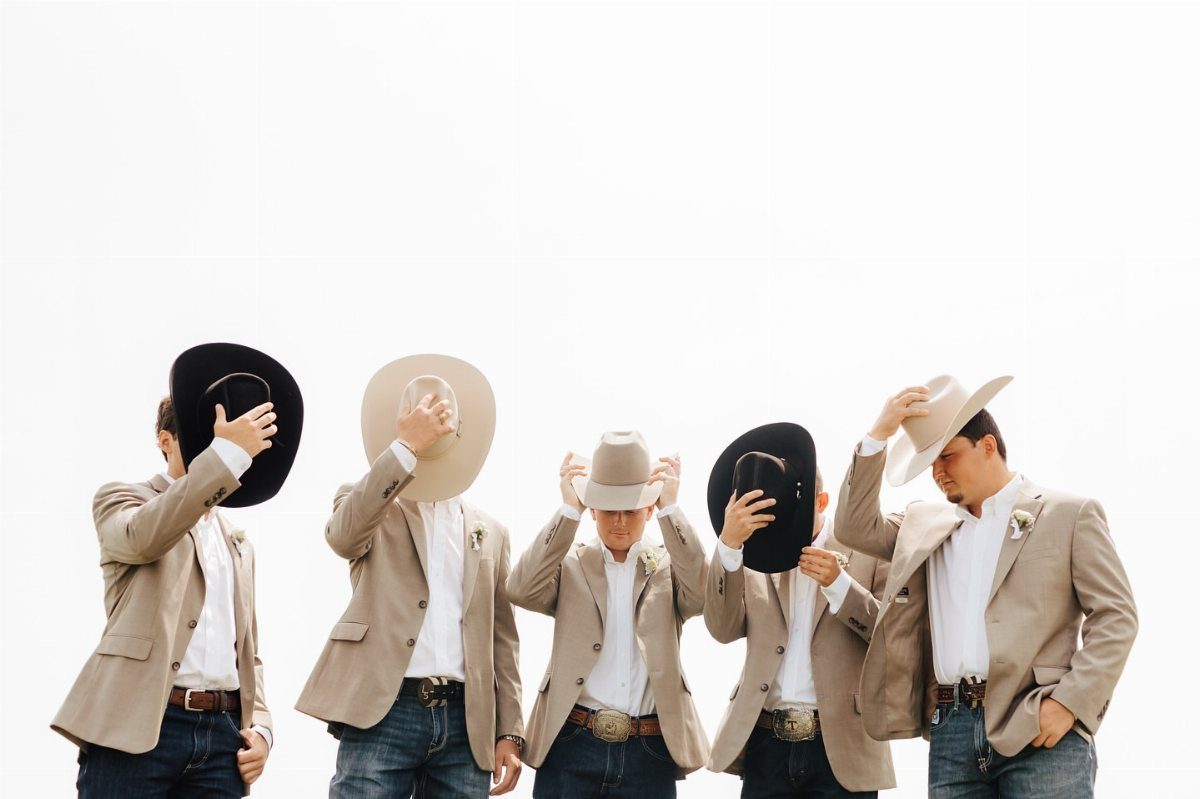 Five groomsmen in tan blazers and cowboy hats posing playfully against white background