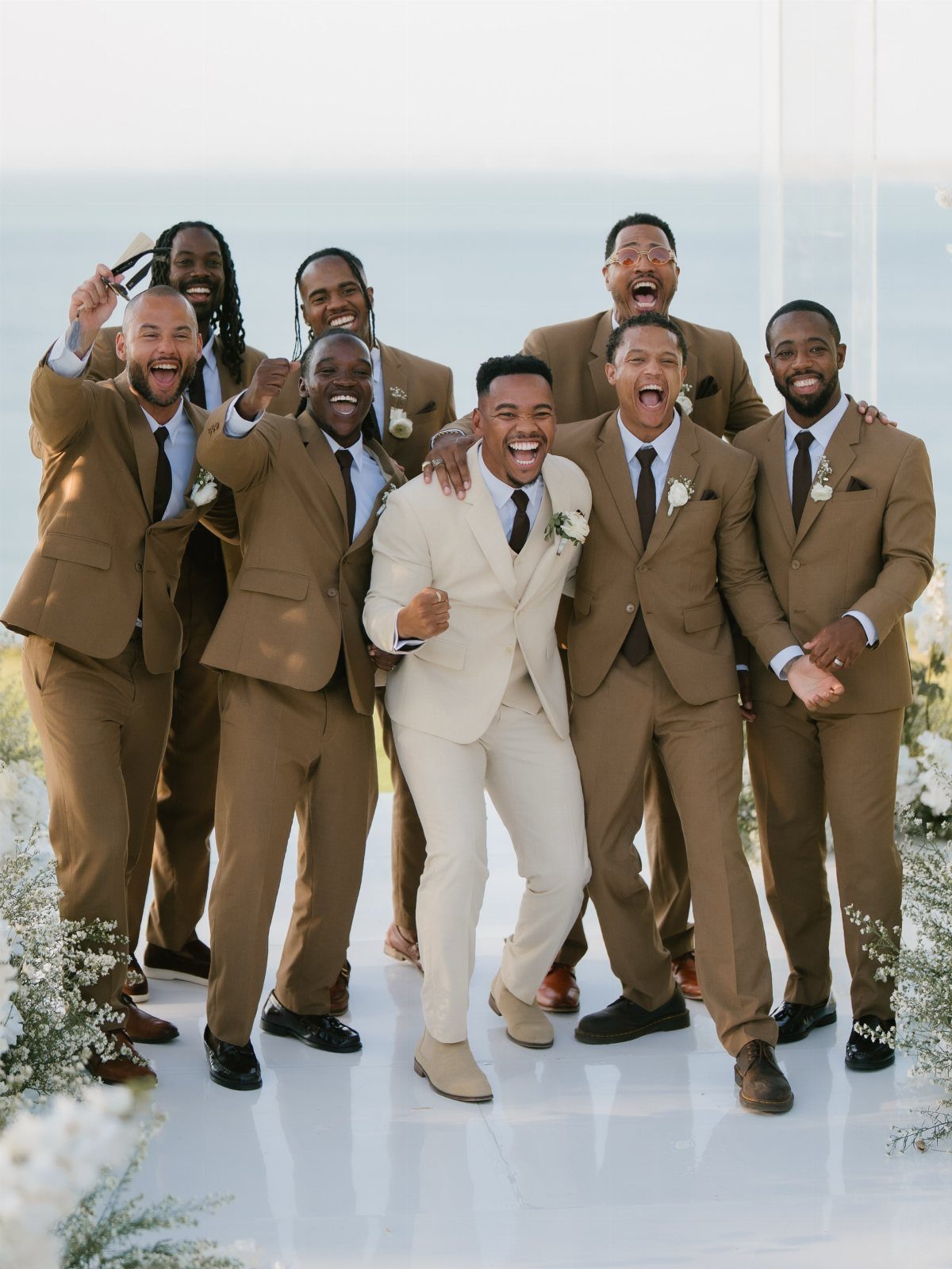 Groom and groomsmen in tan suits cheering with excited expressions by the ocean.
