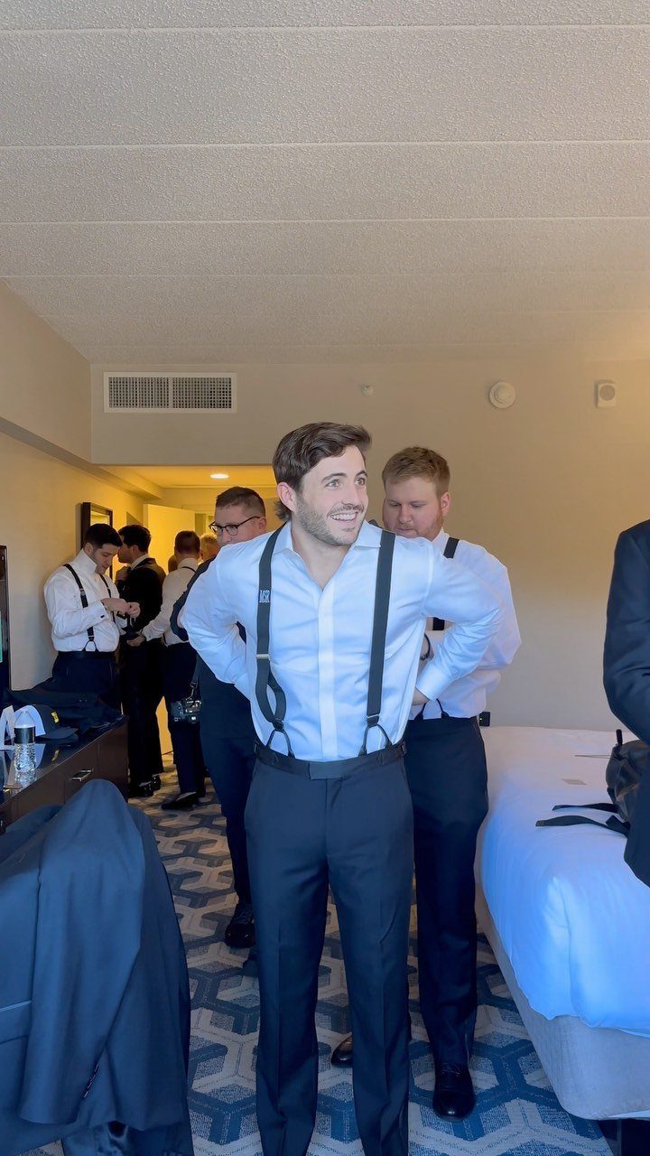 Groom and groomsmen wearing white shirts and suspenders in a hotel room.