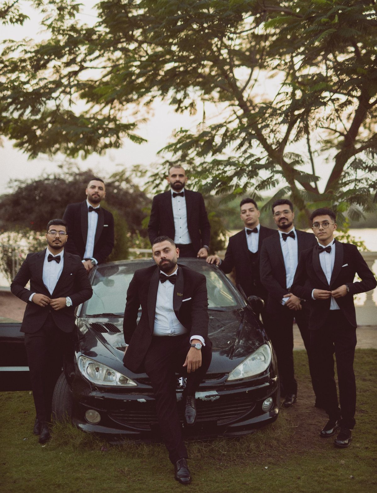 Groomsmen in black tuxedos posing around a black car under a large tree.