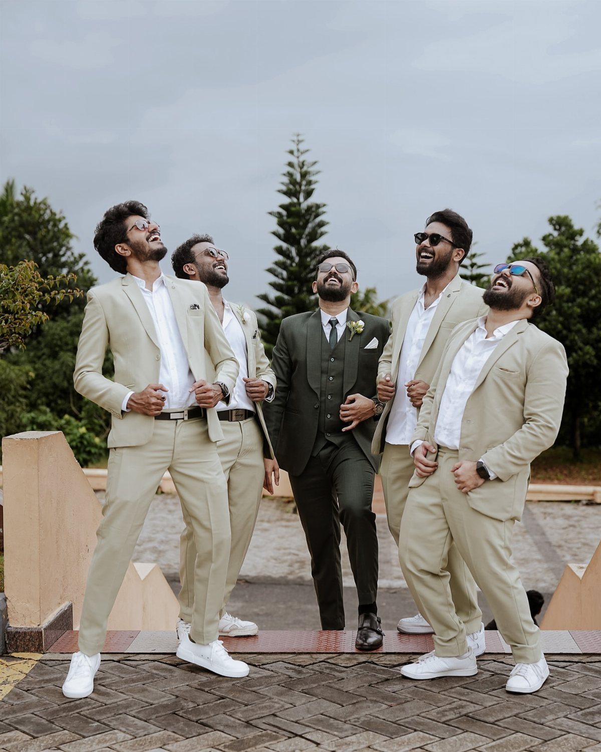 Five men in beige and green suits laughing and looking up outdoors.