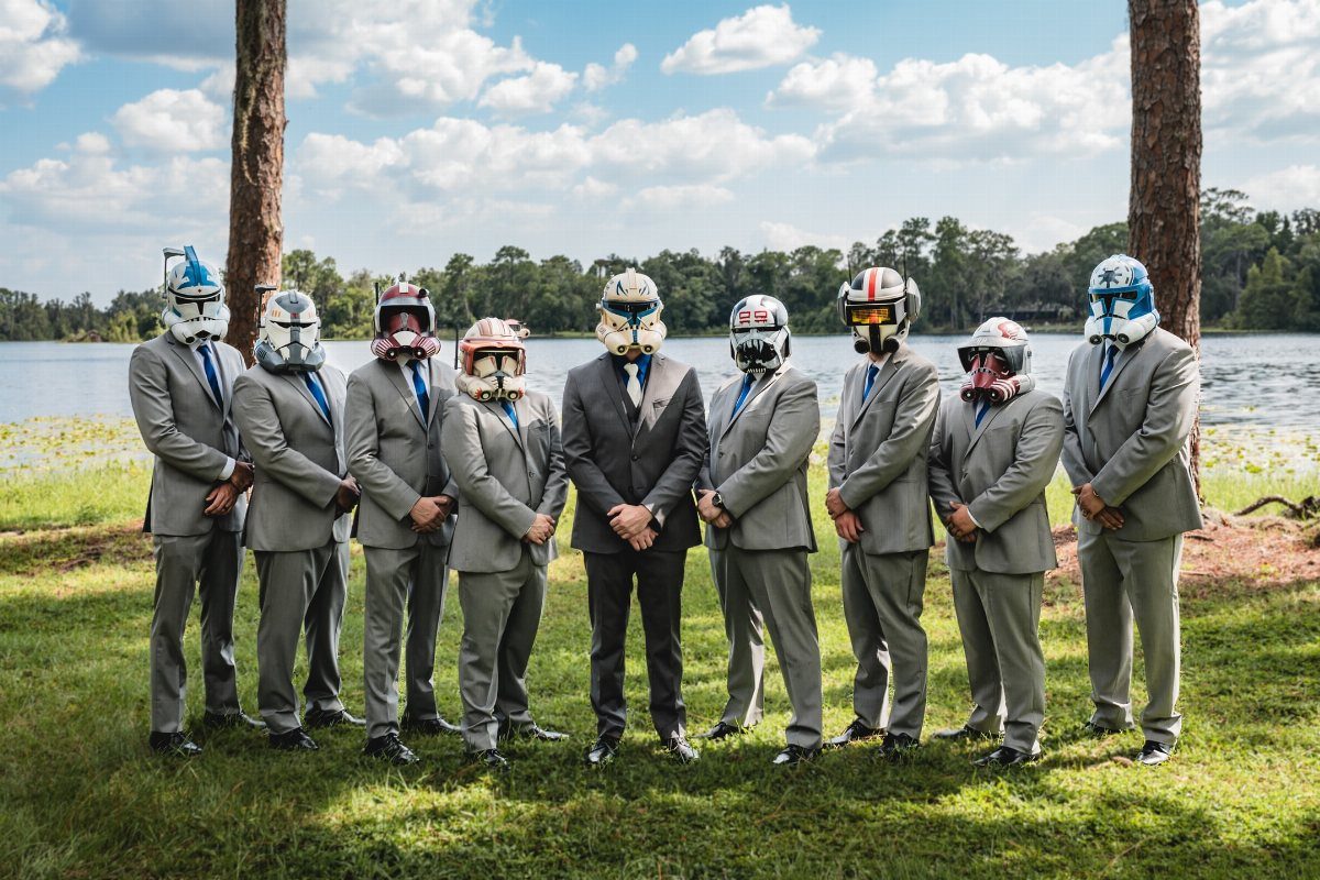 Groomsmen in grey suits wearing football helmets posing by a lake and trees.