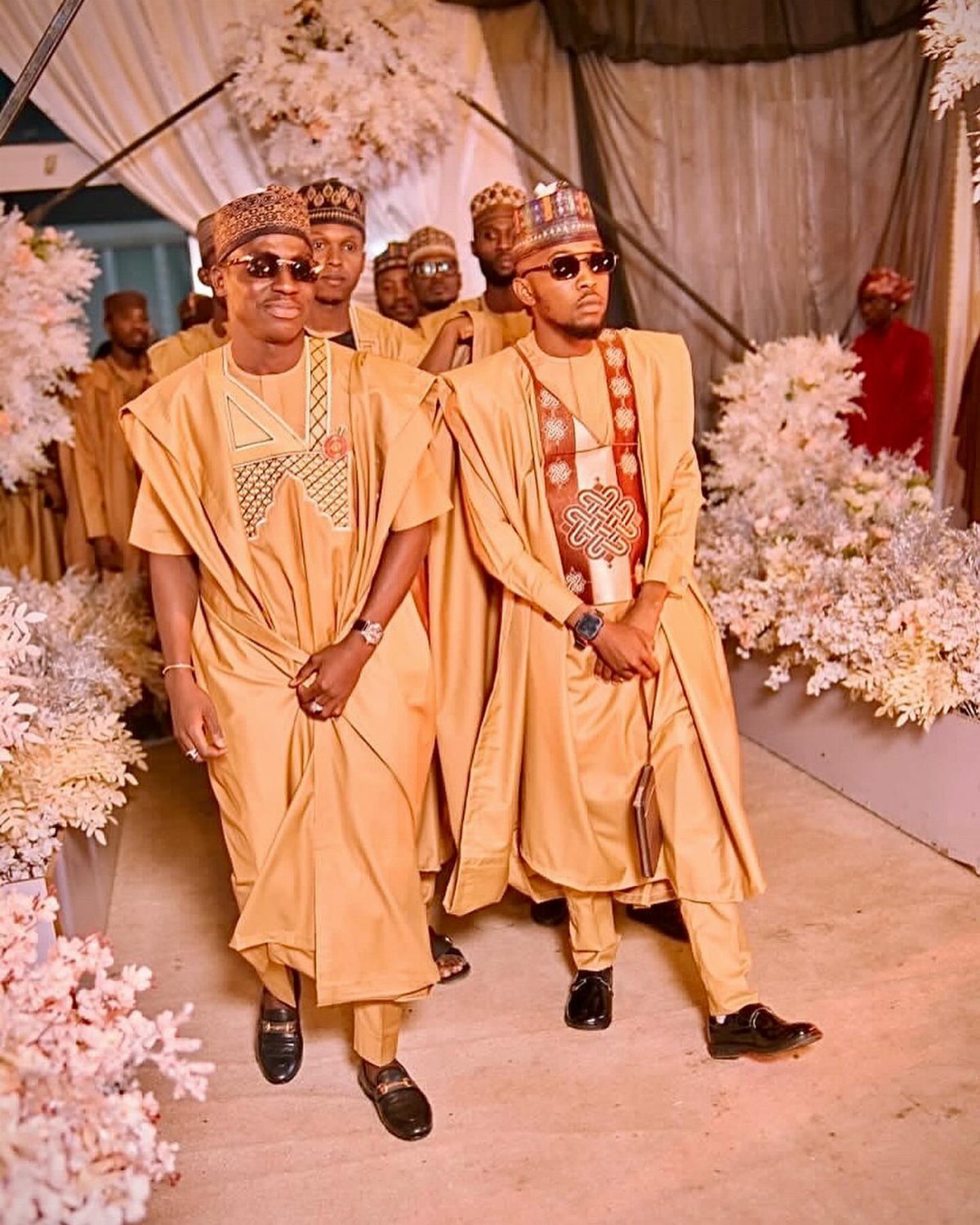 Men in gold traditional Agbada robes walking forward through white floral decorations.