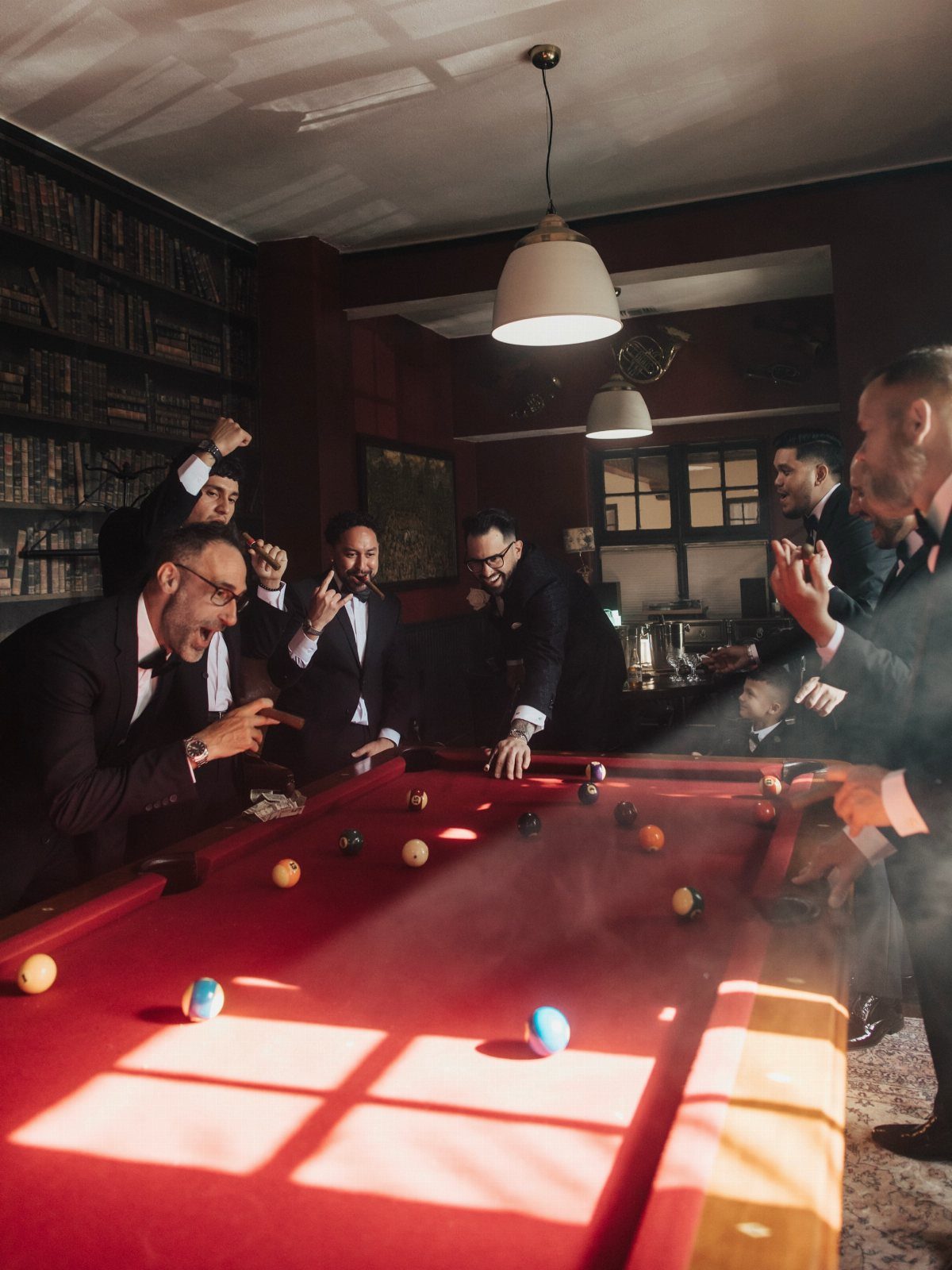 Groomsmen in black tuxedos laughing and playing pool around a red table.