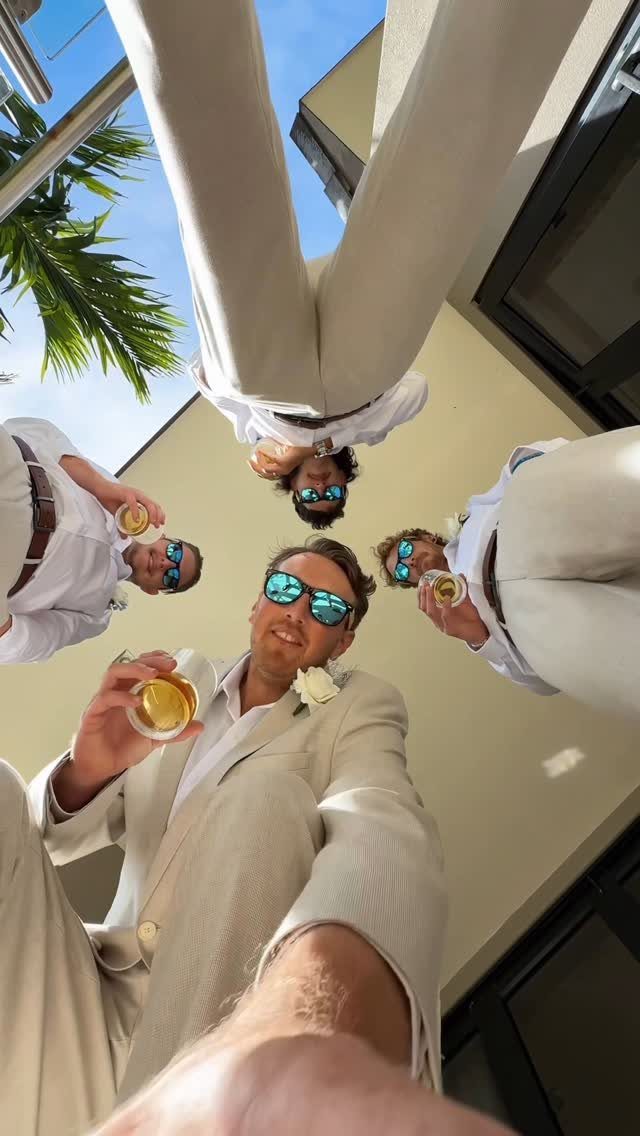 Four men in light suits and sunglasses looking down into a selfie camera.