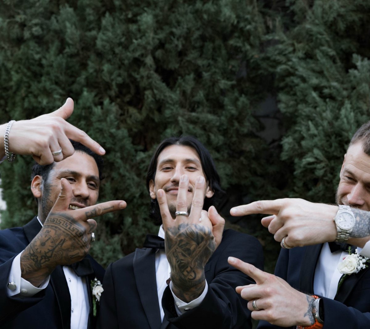 Three groomsmen in tuxedos pointing at the groom's wedding ring outdoors.