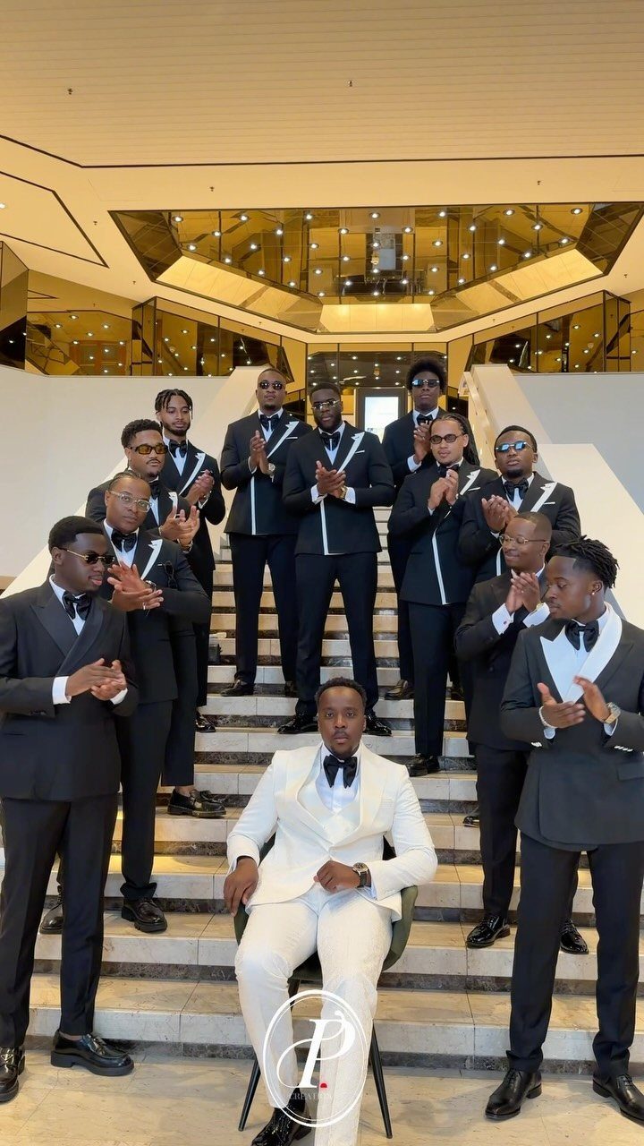 Groom in white tuxedo sitting on stairs surrounded by groomsmen in V formation.