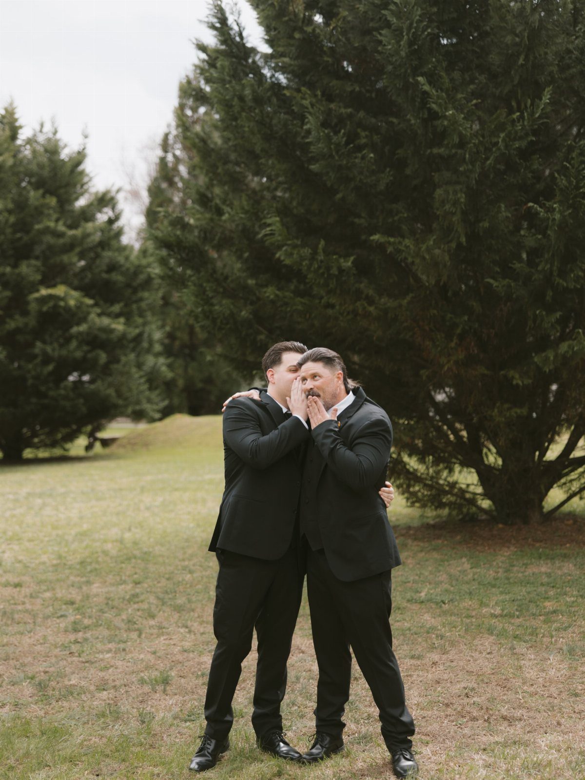 Two men in black suits posing playfully on a grassy field near trees.