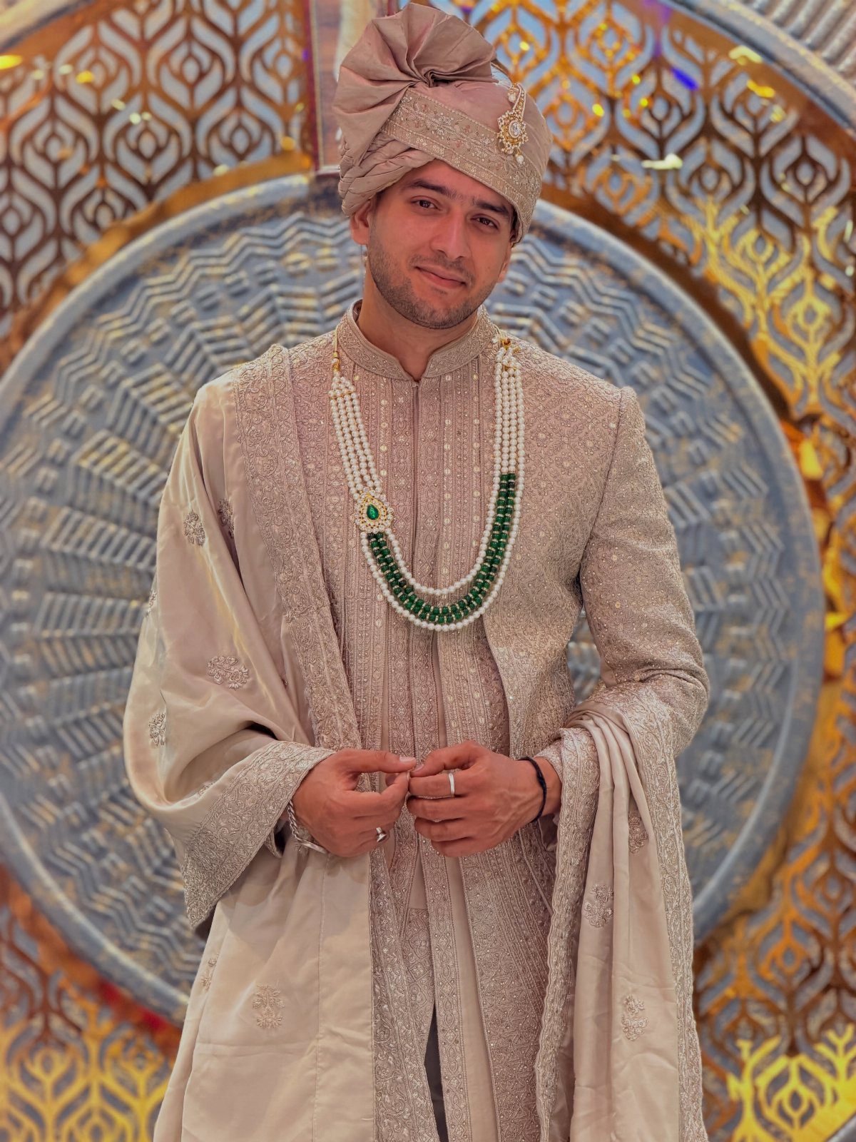 Man in a beige embroidered Indian wedding sherwani with turban and emerald necklace.